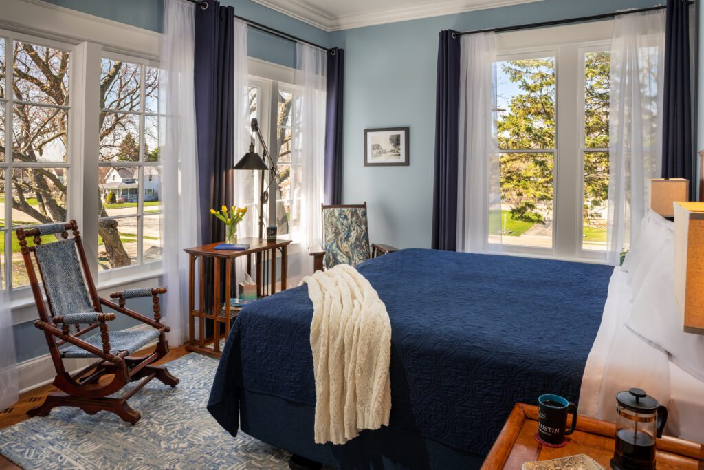 Port_Austin_Bed_and_Breakfast_Port_Austin_Hotel_Room 2-01_Md - Port Austin Bed & Breakfast Image of king bed with blue bedspread with exterior view of trees through picture windows