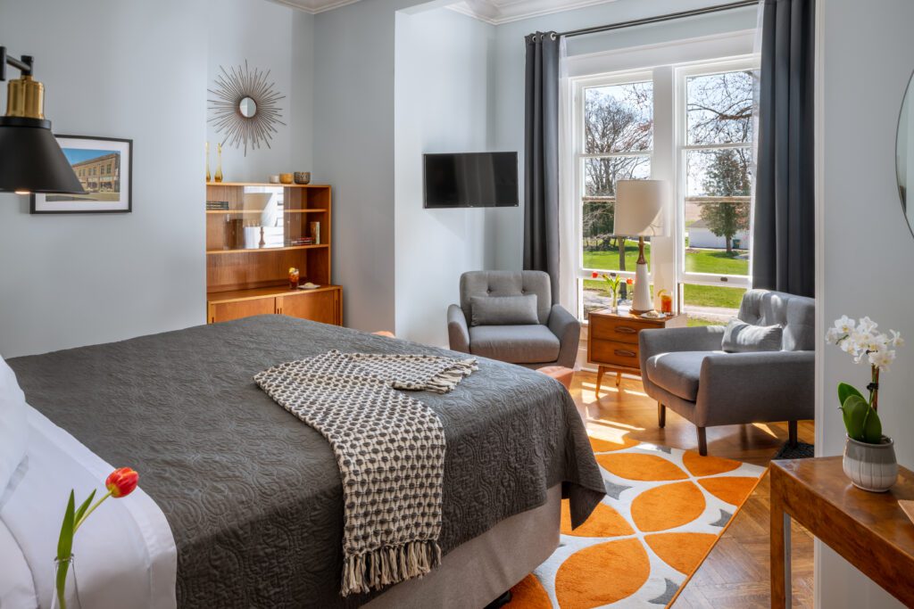 Port_Austin_Bed_and_Breakfast_Port_Austin_Hotel_Room 4-01_Md - Port Austin Bed & Breakfast Image of king bed with gray bedspread and big picture window with tow sitting chairs in front.