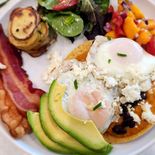 image of huevos rancheros with mango salsa, avocado, over easy eggs, bacon, potato stacks and salad
